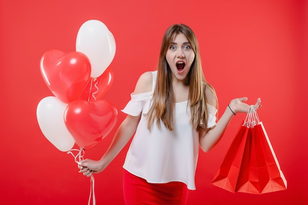 Excited woman with shopping bags and heart shaped balloons isolated over red wall