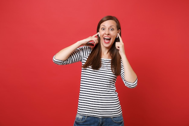 무선 이어폰을 낀 흥분한 여성이 빨간색 배경에 격리된 휴대전화를 들고 놀란 표정으로 입을 크게 벌리고 있습니다. 사람들은 진실한 감정, 라이프 스타일. 복사 공간을 비웃습니다.