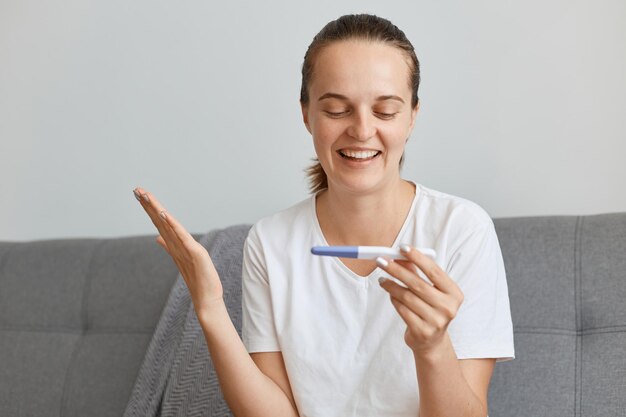 Excited young adult female with pigtail holding positive pregnancy test, looking result with happy optimistic facial expression, raising hand, being happy to become mother.