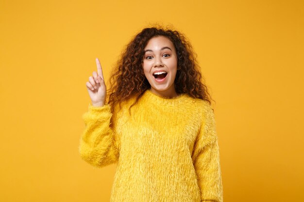 Excited young african american girl in fur sweater posing isolated on yellow orange background, studio portrait. People lifestyle concept. Mock up copy space. Hold index finger up with great new idea.