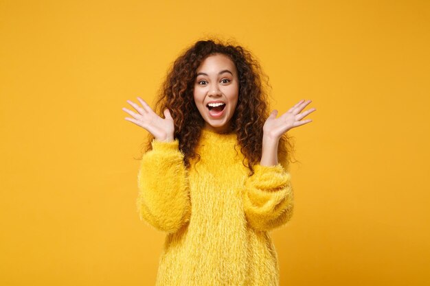 Excited young african american girl in fur sweater posing isolated on yellow orange wall background, studio portrait. People lifestyle concept. Mock up copy space. Keeping mouth open, spreading hands.