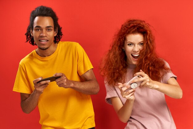 Excited young african-american man and redhead woman playing together and competing in video games on smartphones, win first place or gain highest score, tilt to one side. Relationship and technology.