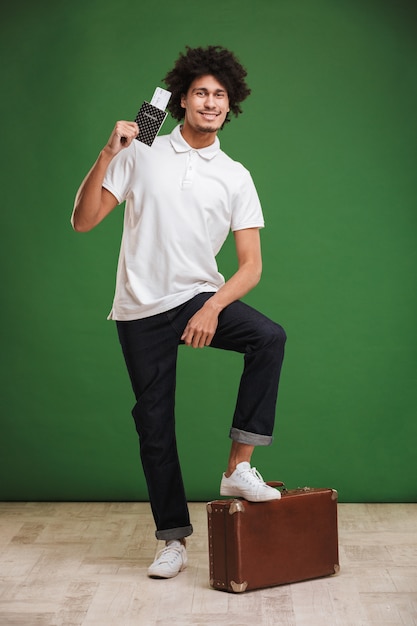 Excited young african curly man holding tickets with passport and suitcase.