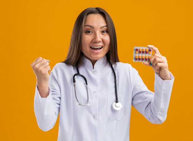 Excited young asian female doctor wearing medical robe and stethoscope showing pack of medical capsules to camera looking at front doing be strong gesture isolated on orange wall