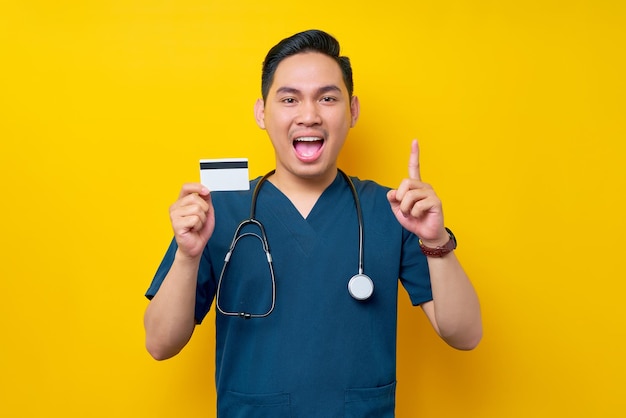 Excited young Asian male doctor or nurse wearing blue uniform holding credit bank card and pointing finger up copy space isolated on yellow background Healthcare medicine concept