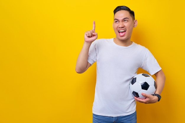 Excited young asian man 20s football fan wearing a white tshirt
carrying a soccer ball and pointing a finger up looking aside
isolated on yellow background people sport leisure lifestyle
concept