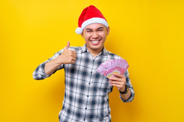 Excited young Asian man 20s wearing a Christmas hat showing cash money rupiah and thumb up sign isolated on yellow background