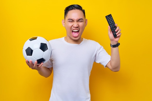 Excited young asian man 20s wearing white tshirt holding a\
soccer ball and remote controller watching the live stream on tv\
isolated on yellow background fans to support football sports\
team