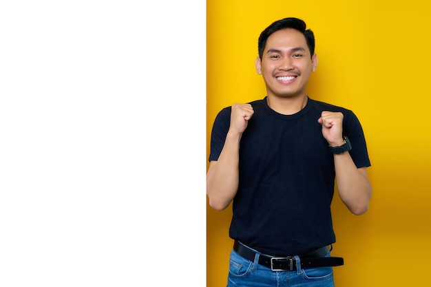 Excited young Asian man in casual tshirt celebrating success with raised fist side of white advertisement board on yellow background Promotion billboard concept