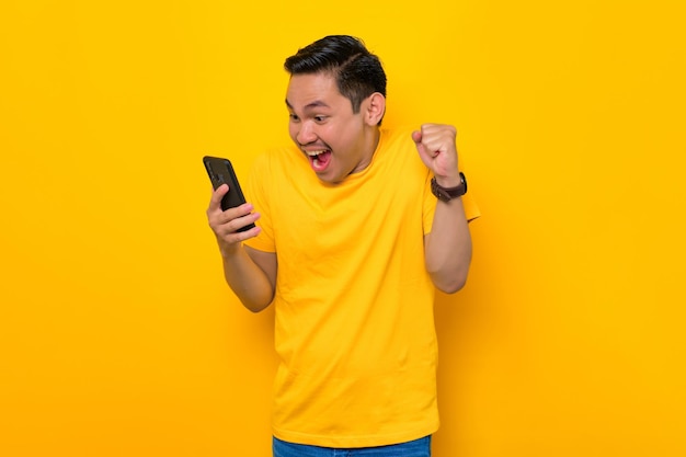 Excited young Asian man in casual tshirt looking at mobile phone feel joyful reading good news isolated on yellow background People lifestyle concept