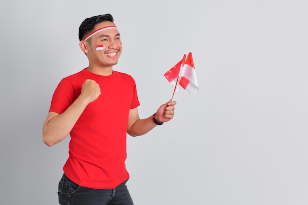 Photo excited young asian man celebrating indonesian independence day holding the indonesian flag isolated on white background