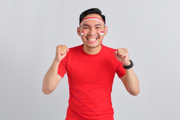 Excited young Asian man celebrating Indonesian independence day standing while raising fist isolated on white background