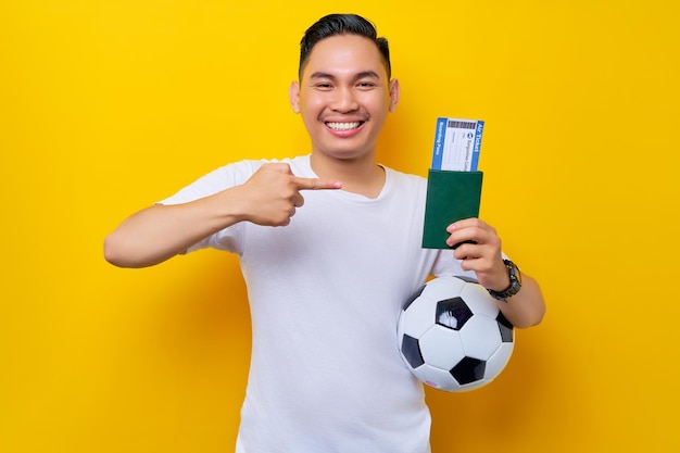 Excited young asian man football fan wearing a white tshirt\
carrying a soccer ball and pointing a finger at a passport ticket\
boarding pass isolated on yellow background