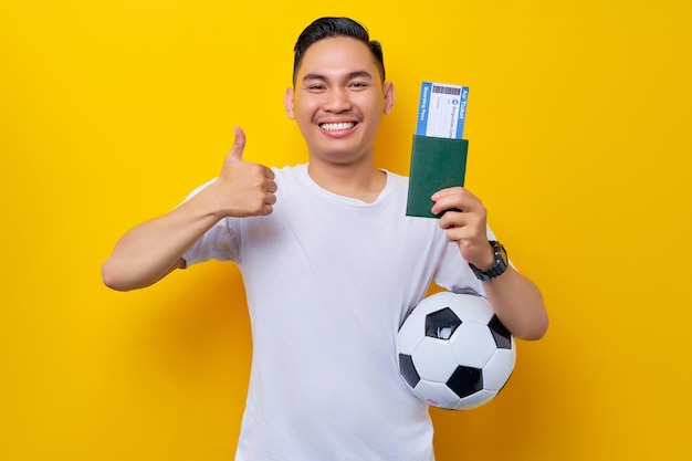 Excited young asian man football fan wearing a white tshirt
carrying a soccer ball and showing passport ticket boarding pass
with thumb up gesture isolated on yellow background