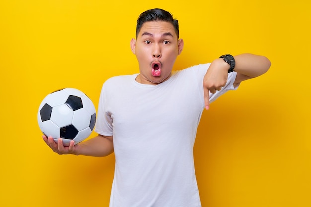 Excited young asian man football fan wearing a white tshirt holding a soccer ball and pointing a finger down on promo isolated on yellow background people sport leisure lifestyle concept