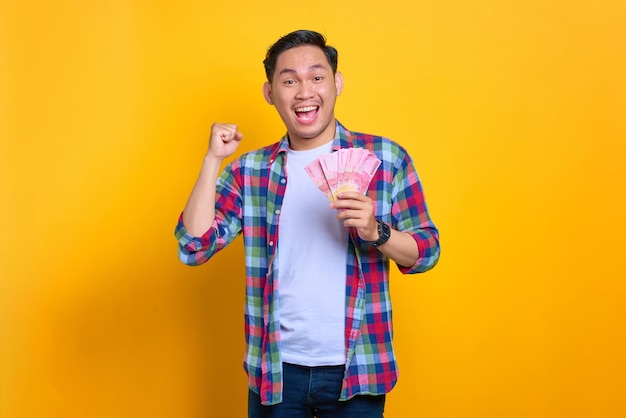 Excited young asian man in plaid shirt holding money banknotes and celebrating success isolated on yellow background