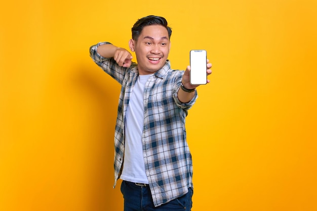 Excited young Asian man in plaid shirt showing mobile phone blank screen recommending app isolated on yellow background