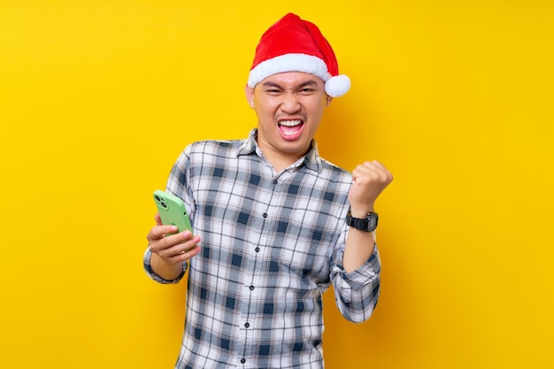クリスマス帽子をかぶった格子縞のシャツを着た興奮した若いアジア人男性が携帯電話を持ち、黄色の背景に「はい」と言って勝者のジェスチャーをしているクリスマスと新年あけましておめでとうございます2023年の休日のコンセプト