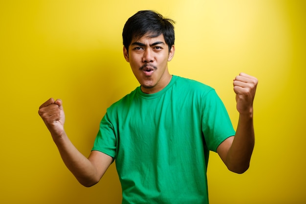 Excited young asian man raising his fists with smiling delighted face, yes gesture, celebrating success on yellow background