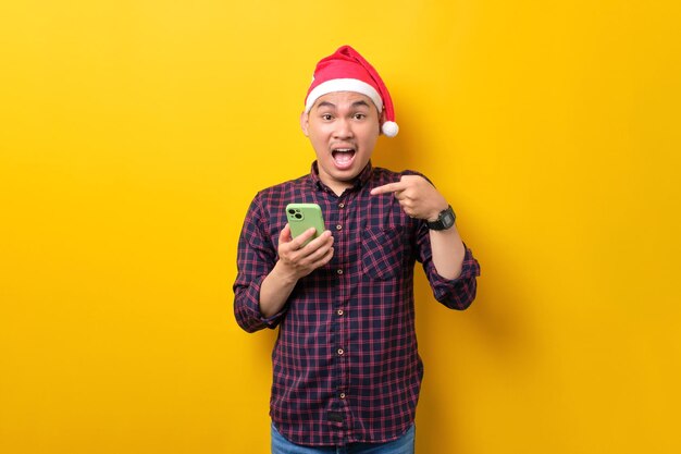 Excited young Asian man in Santa hat pointing finger at smartphone recommending mobile application on yellow studio background Happy New Year 2023 celebration merry holiday concept