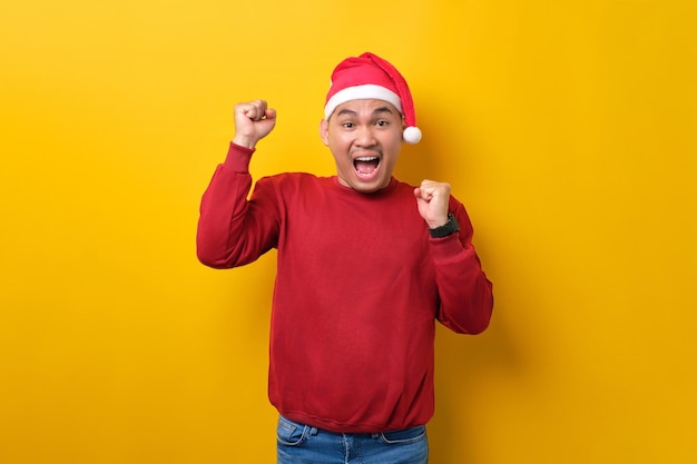 Excited young Asian man in Santa hat raising hands up celebrating success on yellow studio background celebration Christmas holiday and New Year concept