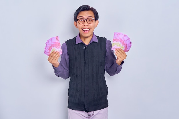 흰색 배경에 격리된 루피아 지폐에 현금 돈을 보여주는 셔츠 옷을 입은 흥분된 젊은 아시아 남자 사람들의 라이프 스타일 개념