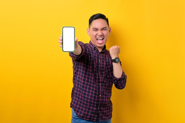 Excited young Asian man showing smartphone with empty screen celebrating win isolated on yellow background Advertising new mobile app mockup concept