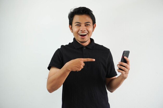 Photo excited young asian man smiling happy while pointing to mobile phone that he hold isolated