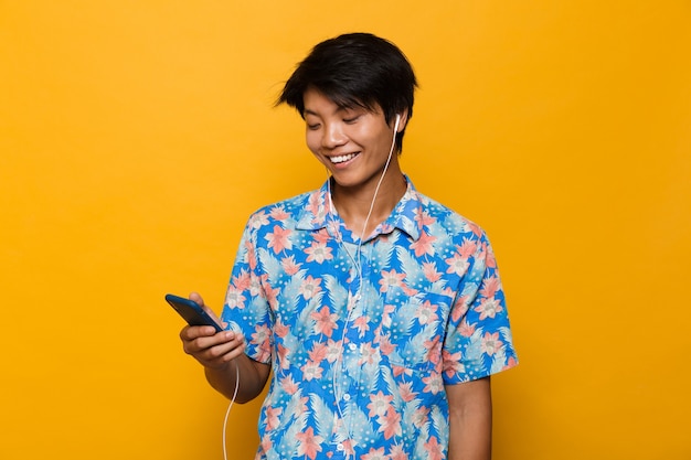 Excited young asian man standing isolated over yellow space using mobile phone listening music with earphones.