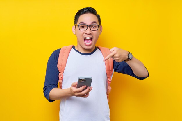 Excited young asian man student in casual clothes and backpack glasses pointing finger at mobile phone recommending app isolated on yellow background high school university college concept