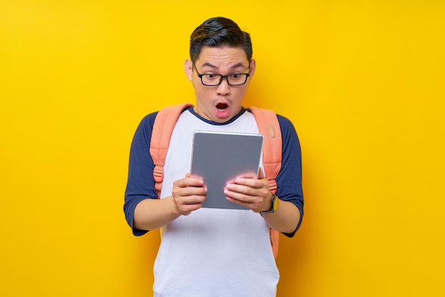 Excited young asian man student in casual clothes and glasses backpack using digital tablet computer isolated on yellow background high school university college concept