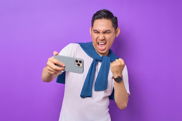 Excited young asian man using smartphone for playing mobile game celebrating success isolated over purple background