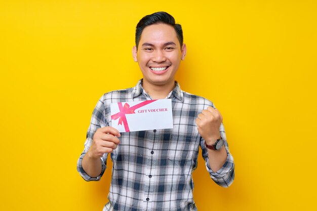 Excited young asian man wearing a white checkered shirt holding a gift voucher and celebrating the winner isolated on yellow background people lifestyle concept