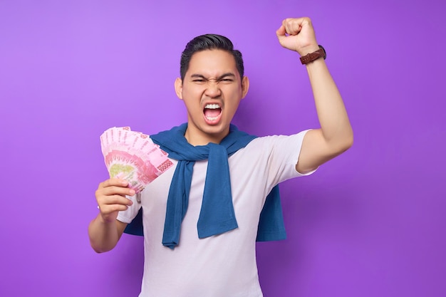 Возбужденный молодой азиат в белой футболке в повседневной одежде держит денежные банкноты и сжимает кулак, празднуя финансовый успех, изолированный на фиолетовом фоне Концепция прибыли и богатства