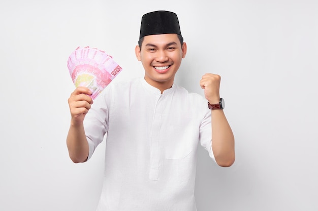 Excited young Asian Muslim man holding cash money rupiah banknotes and celebrating success isolated on white background People religious Islamic lifestyle concept