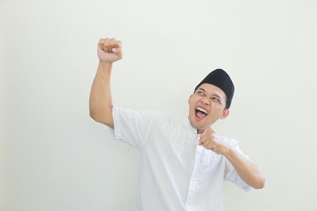 Excited young Asian Muslim man raising fists and celebrating victory