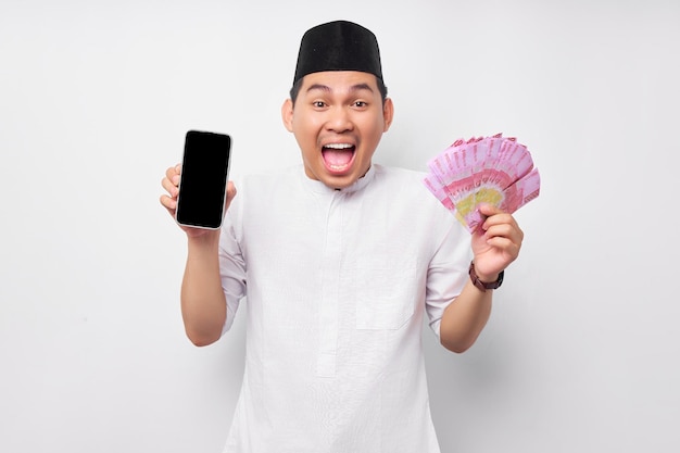 Excited young Asian Muslim man showing mobile phone with blank screen and cash money rupiah banknotes isolated on white background People religious Islamic lifestyle concept