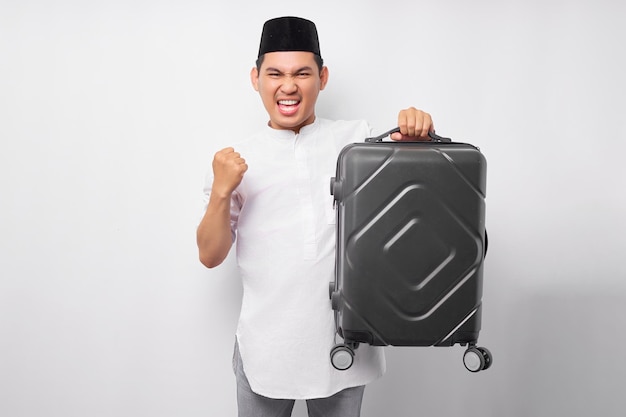 Excited young Asian Muslim man standing while holding a suitcase in hand celebrating success and ready for the holiday eid Mubarak isolated on white background Ramadan and eid Mubarak concept