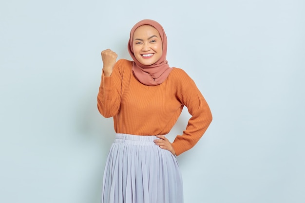갈색 스웨터를 입은 젊은 아시아 무슬림 여성이 흰색 배경에 고립되어 있다고 말하는 주먹을 축하하는 승리 제스처를 하고 있다