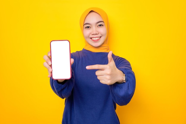 Excited young Asian Muslim woman dressed in casual sweater showing blank screen mobile phone recommending mobile app isolated over yellow background