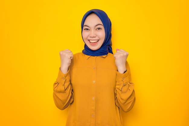 Excited young asian muslim woman dressed in orange celebrating success with raised fists isolated on yellow background