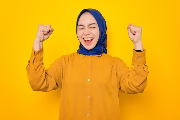 Excited young Asian Muslim woman dressed in orange shirt celebrating luck or success with raised fists isolated on yellow background