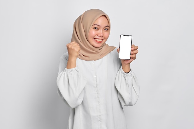 白い背景の上に分離されたアプリをお勧めする携帯電話の空白の画面を示す興奮した若いアジアのイスラム教徒の女性