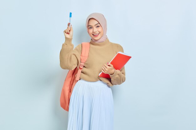 Excited young Asian Muslim woman student in brown sweater with backpack holding book and having good idea isolated on white background back to school concept