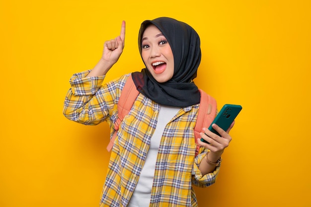 Excited young Asian Muslim woman student in Plaid shirt and backpack holding mobile phone pointing finger up creating genius solutions isolated on yellow background