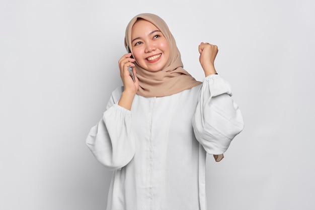 Excited young Asian Muslim woman talking on mobile phone and doing winner gesture isolated over white background