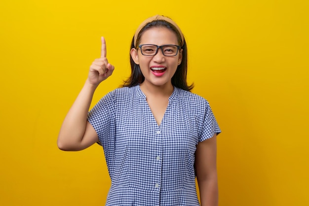 Excited young asian woman 30s wearing a dress checkered with\
glasses looking smart and pointing her finger up asking questions\
having creative ideas on yellow background people lifestyle\
concept