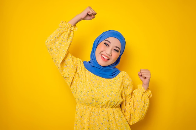 Возбужденная молодая азиатка в повседневной одежде празднует победу и поднимает кулаки на желтом фоне