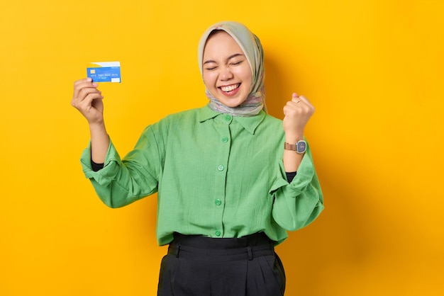 Excited young Asian woman in a green shirt holding credit card and makes winner gesture on yellow background