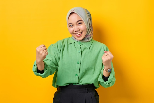 Excited young Asian woman in green shirt raised hands celebrating success on yellow background